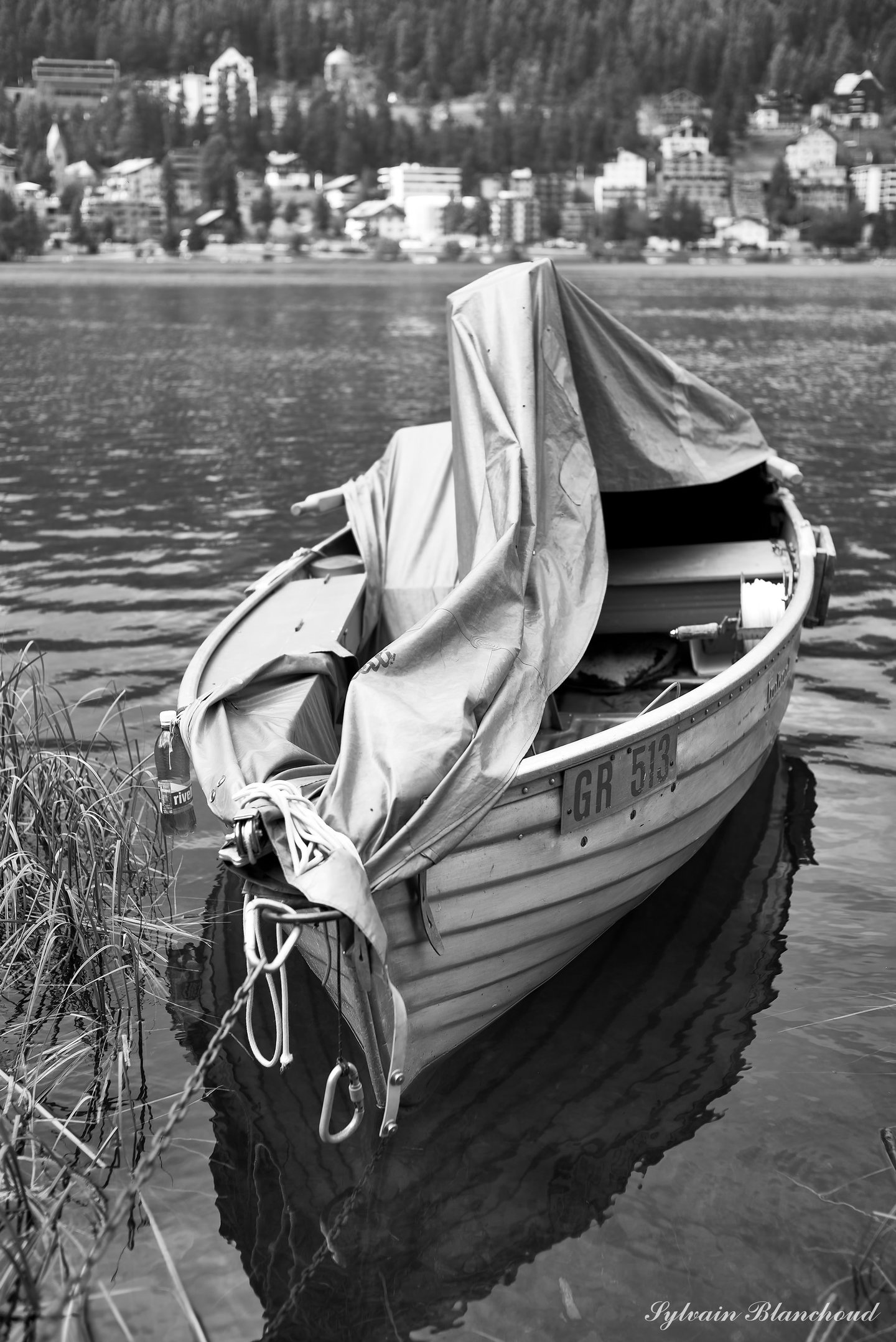 Noir et blanc : une barque en bois couverte d'une bâche froissée, amarrée dans les roseaux ; une ville s'étage sur la colline au loin.