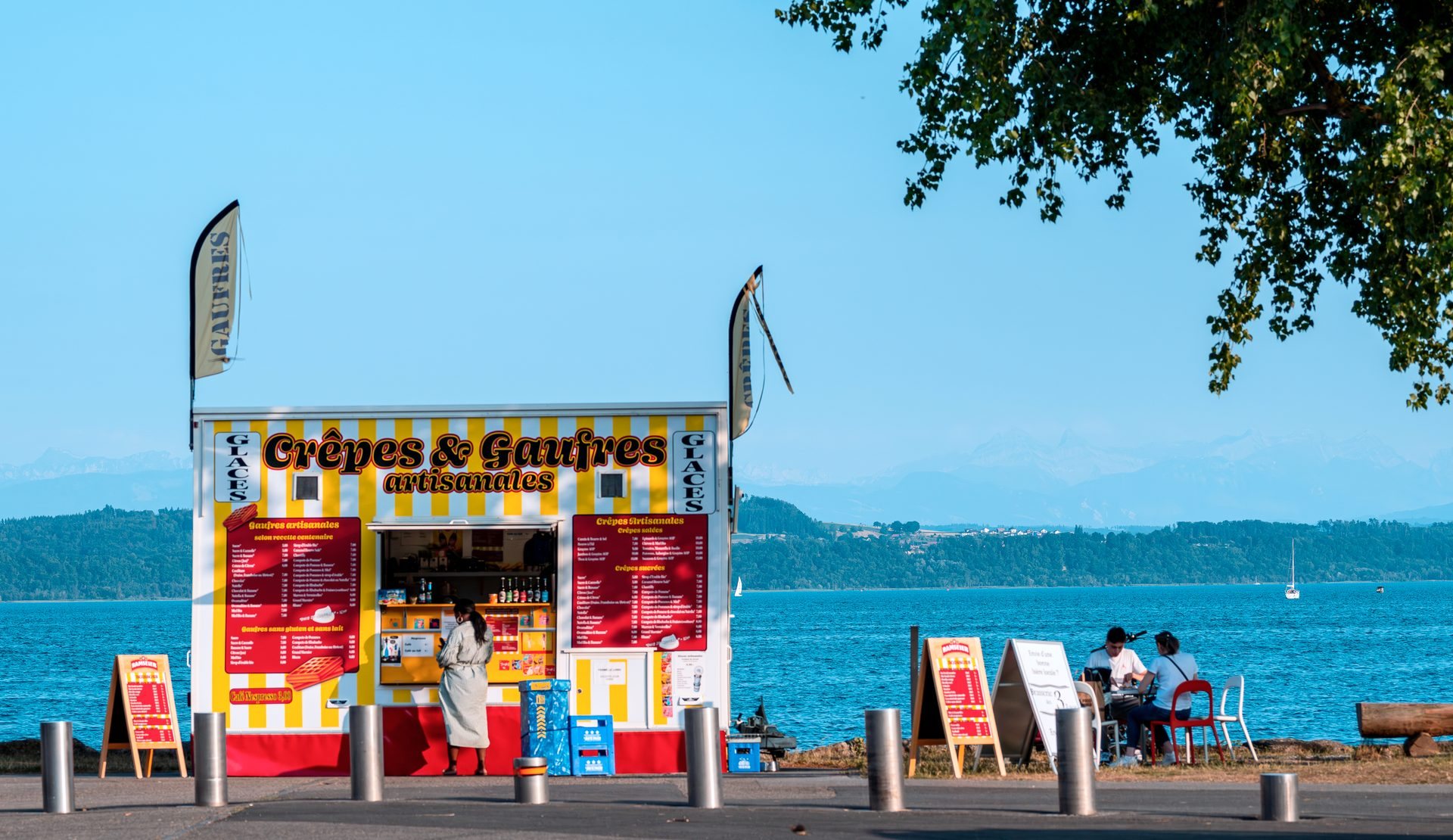 Un kiosque à crêpes et gaufres aux rayures rouges et jaunes, au bord du lac de Neuchâtel, sous un ciel bleu ; deux promeneurs sont attablés à côté.