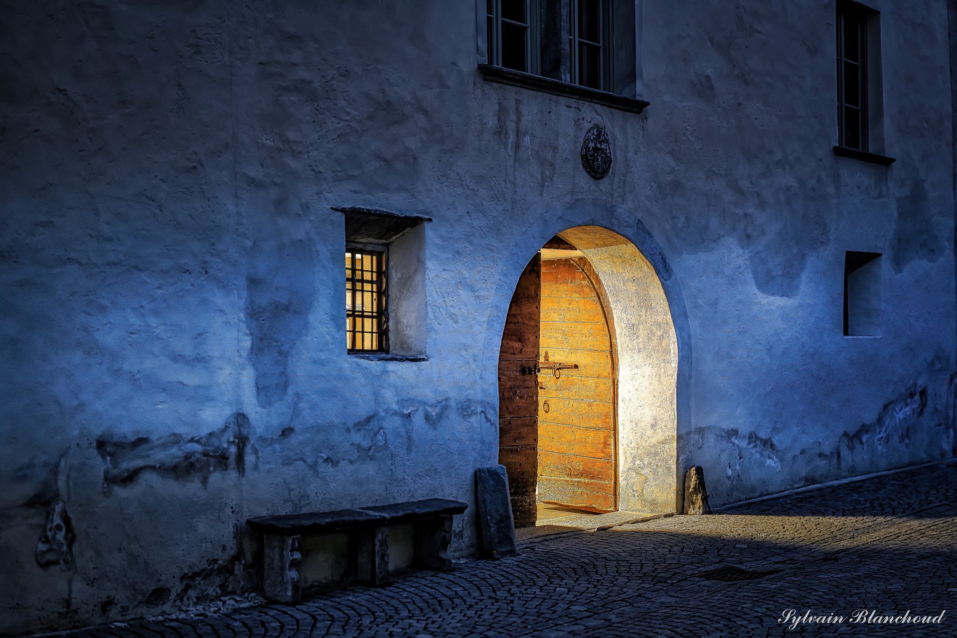 Une façade crépie baignée d'une lumière bleue, une porte de bois cintrée éclairée d'or et un banc de pierre au pied du mur.