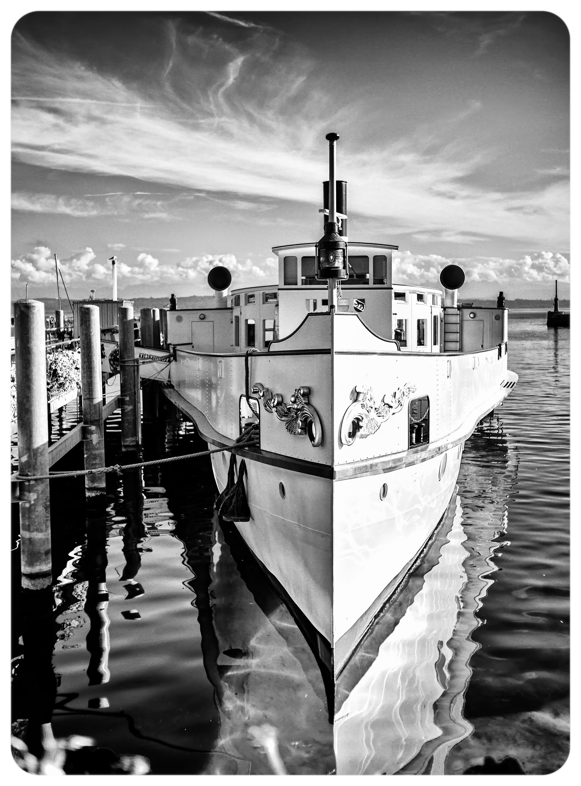 Noir et blanc : un bateau à vapeur blanc amarré au ponton, vu de face, son reflet tremblant dans l'eau calme du port.