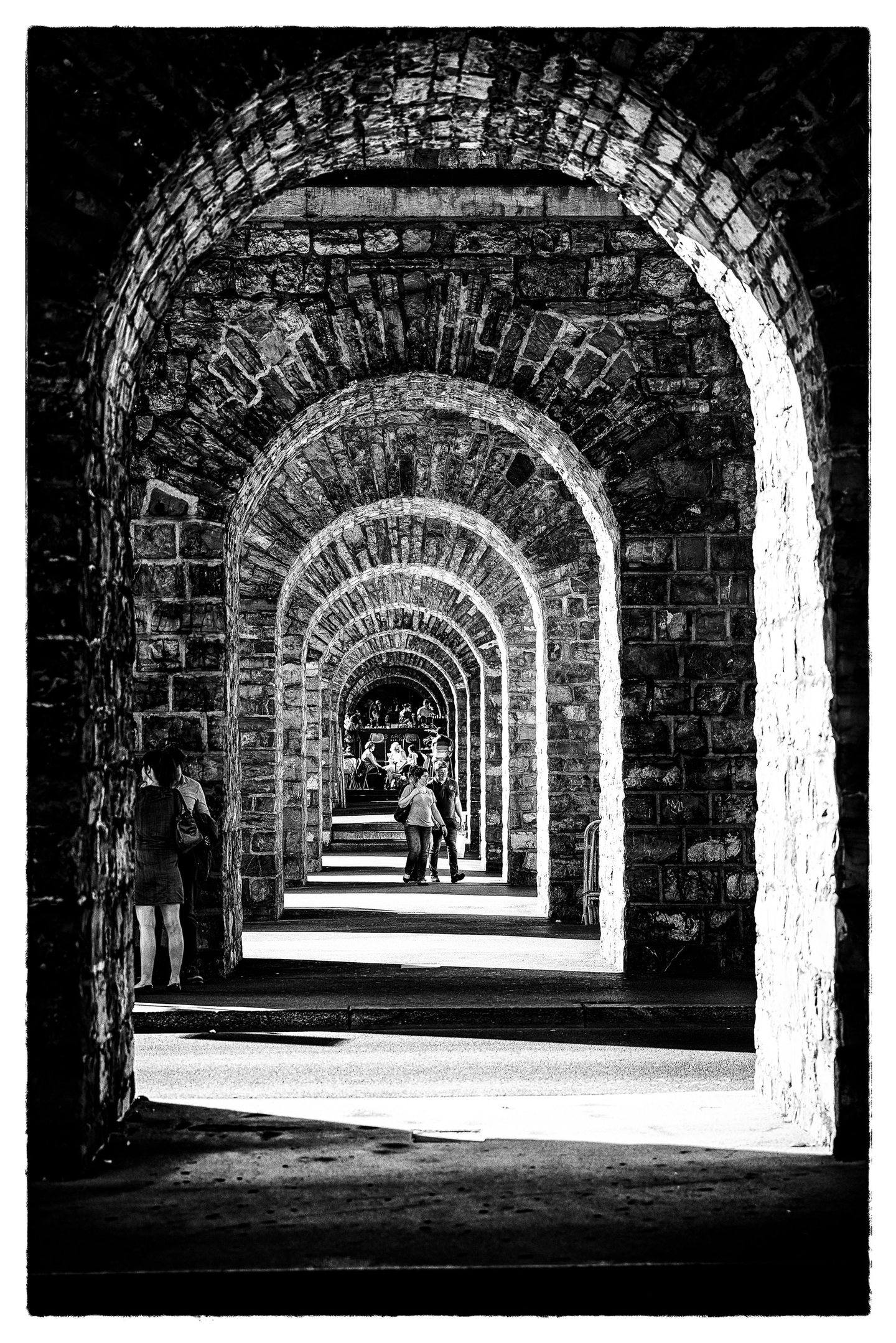 Noir et blanc : une enfilade d'arches de pierre qui se répètent jusqu'à un point de lumière ; quelques silhouettes marchent au loin.