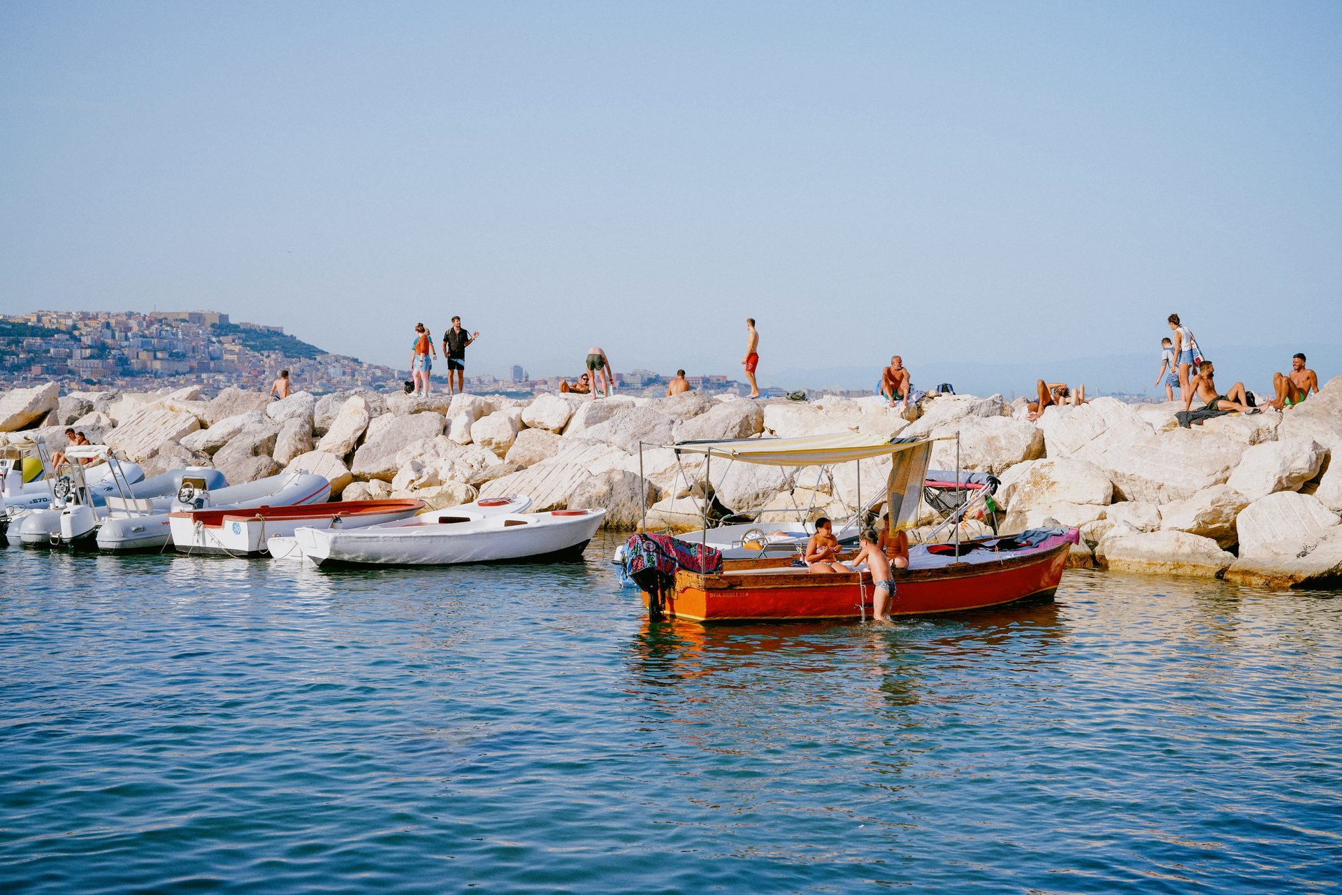 Une barque rouge devant une digue de rochers blancs où des baigneurs prennent le soleil, sur une mer d'un bleu limpide.