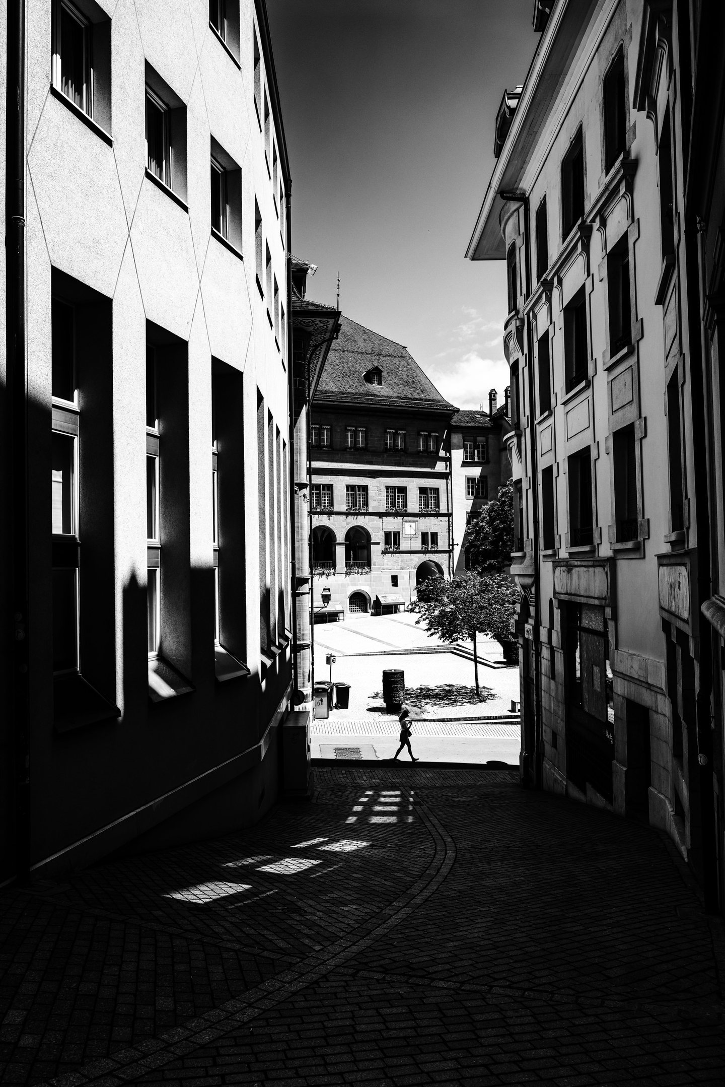 Noir et blanc : une ruelle étroite entre de hautes façades ; au bout, une place inondée de soleil où passe une silhouette.