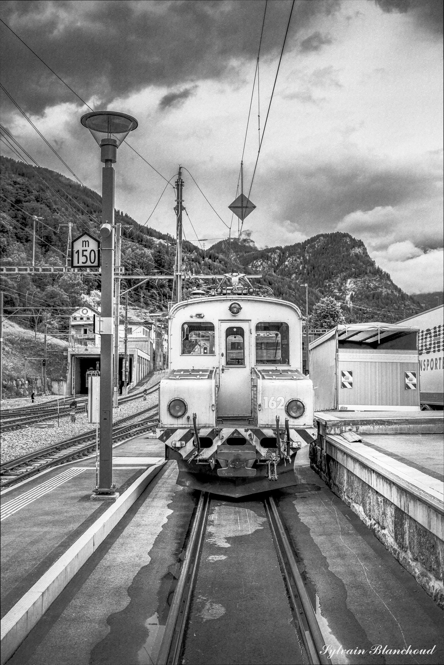 Noir et blanc : un ancien train à l'arrêt au bout de la voie, sous les caténaires, des montagnes et un ciel chargé derrière lui.